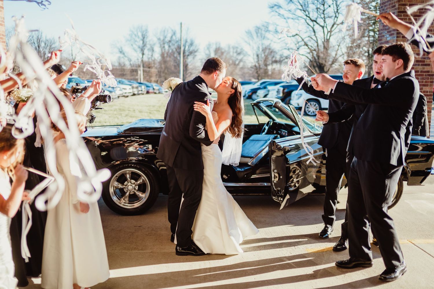 Bride and groom kissing during wedding send-off with guests waving ribbon wands by a classic black car. Indianapolis wedding photographer