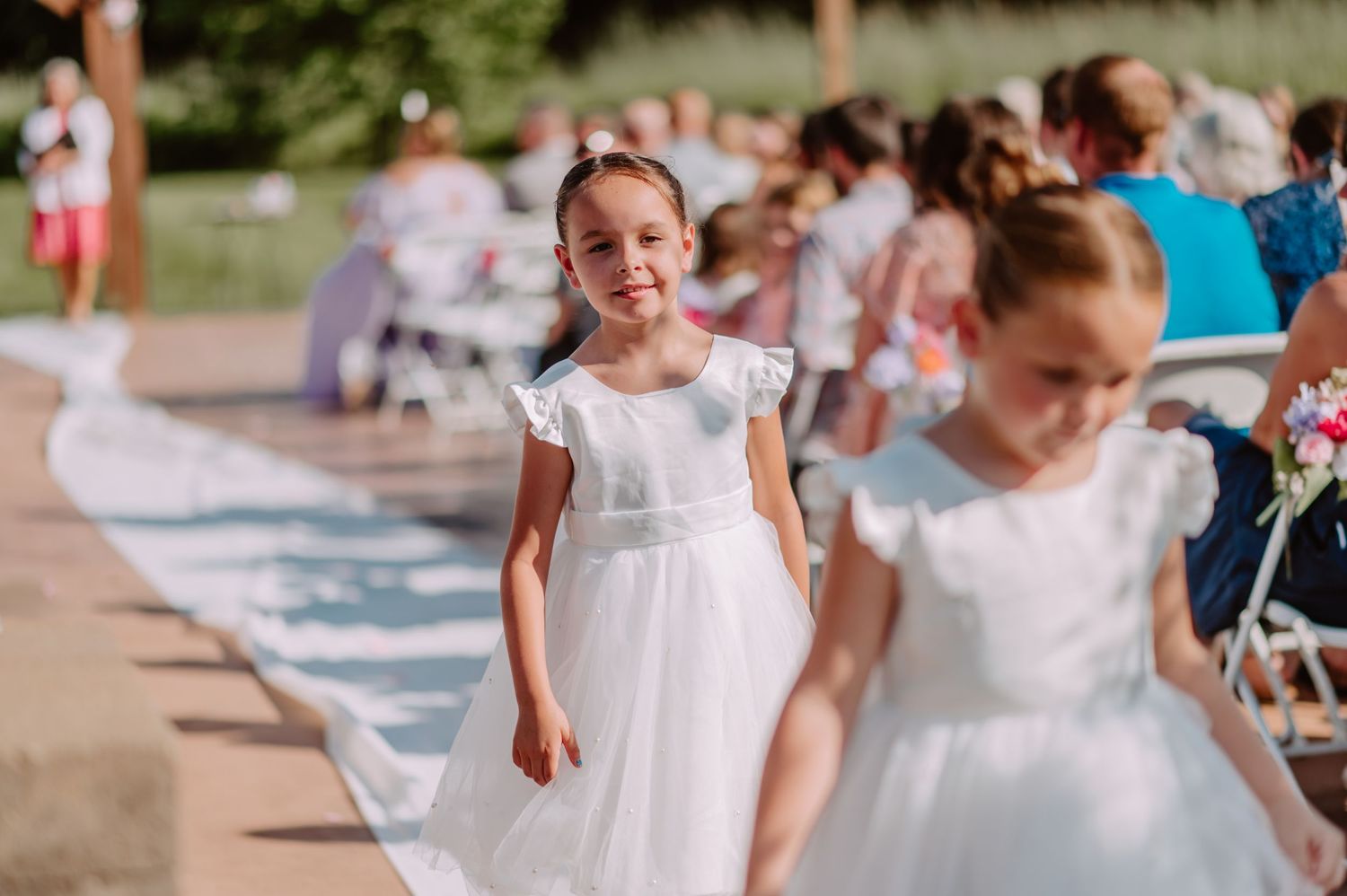 Sweet flower girl in white dress smiles while walking down the outdoor wedding ceremony aisle.