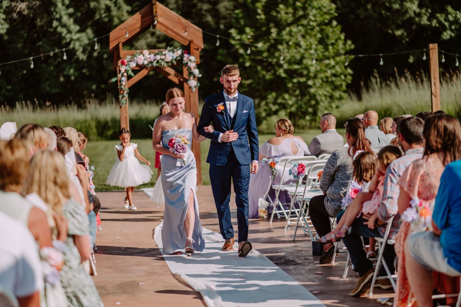Groom in navy suit walks down outdoor wedding aisle with bridesmaid in blue dress, guests seated on both sides.