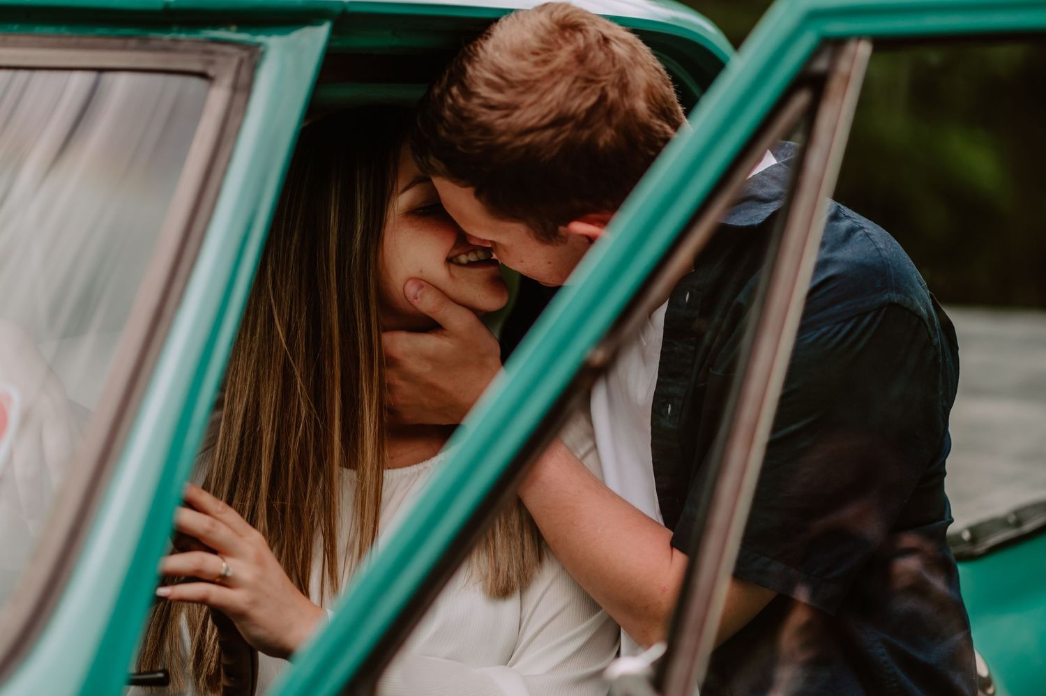 Romantic couple sharing a kiss leaning through the window of a vintage teal green car outdoors.