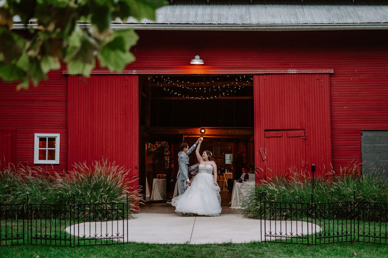 Bride in white gown stands in doorway of red barn wedding venue with string lights and lush greenery. Indianapolis wedding photographer