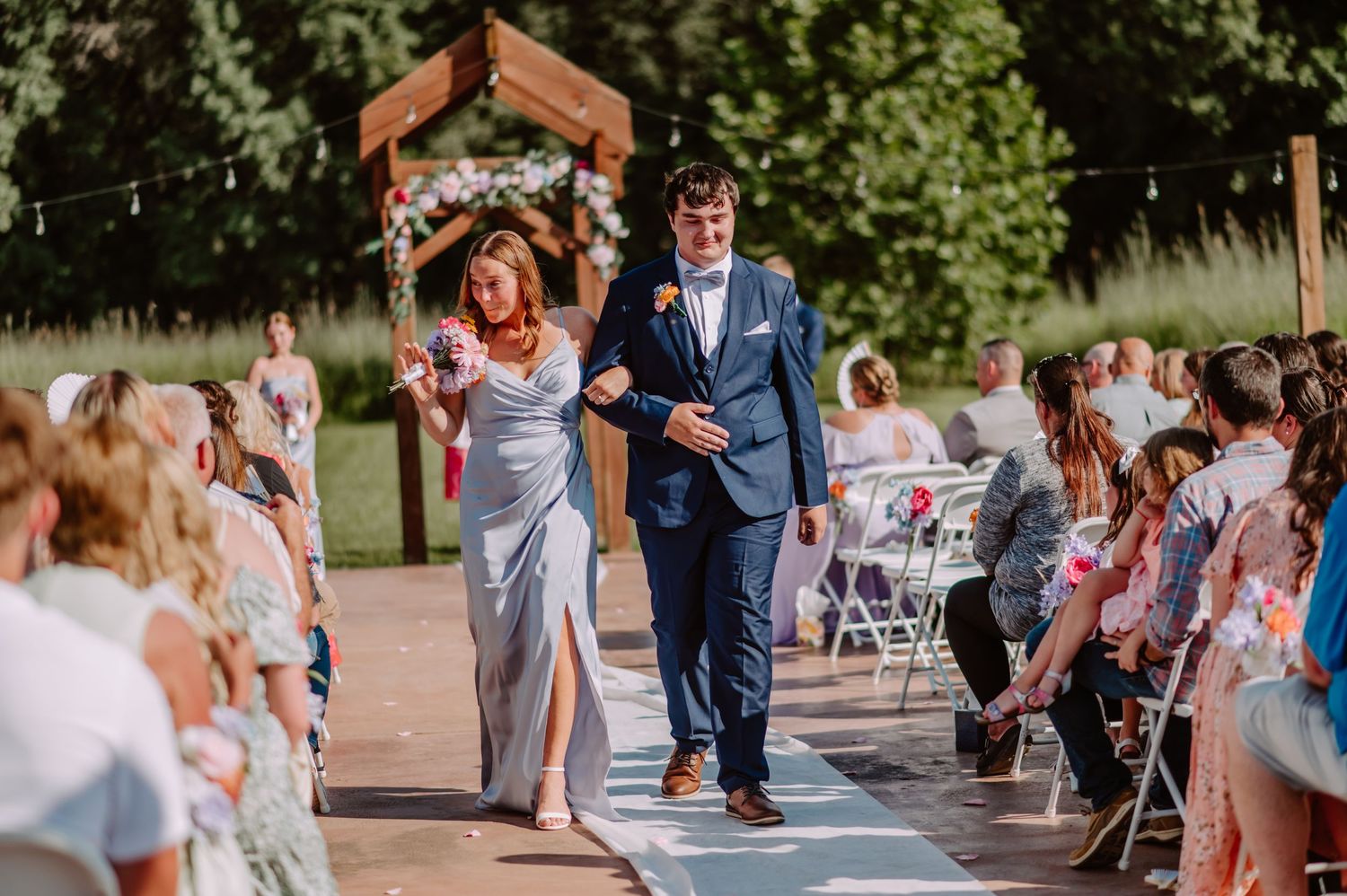 Bridesmaid in silver-blue dress walks arm-in-arm with groomsman in navy suit down outdoor wedding aisle.