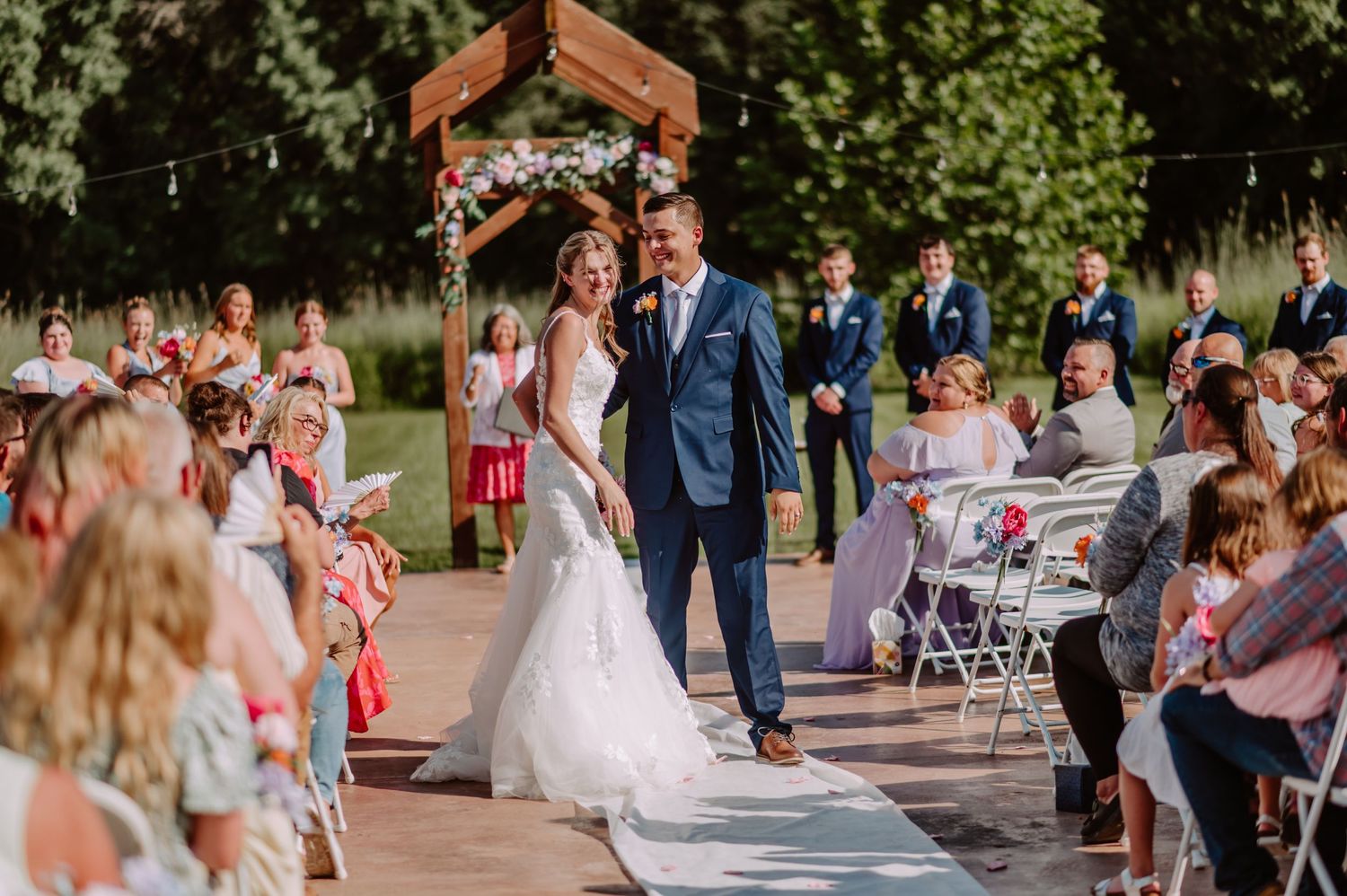 Joyful bride and groom walk down outdoor wedding aisle surrounded by seated guests and wooden floral arch.
