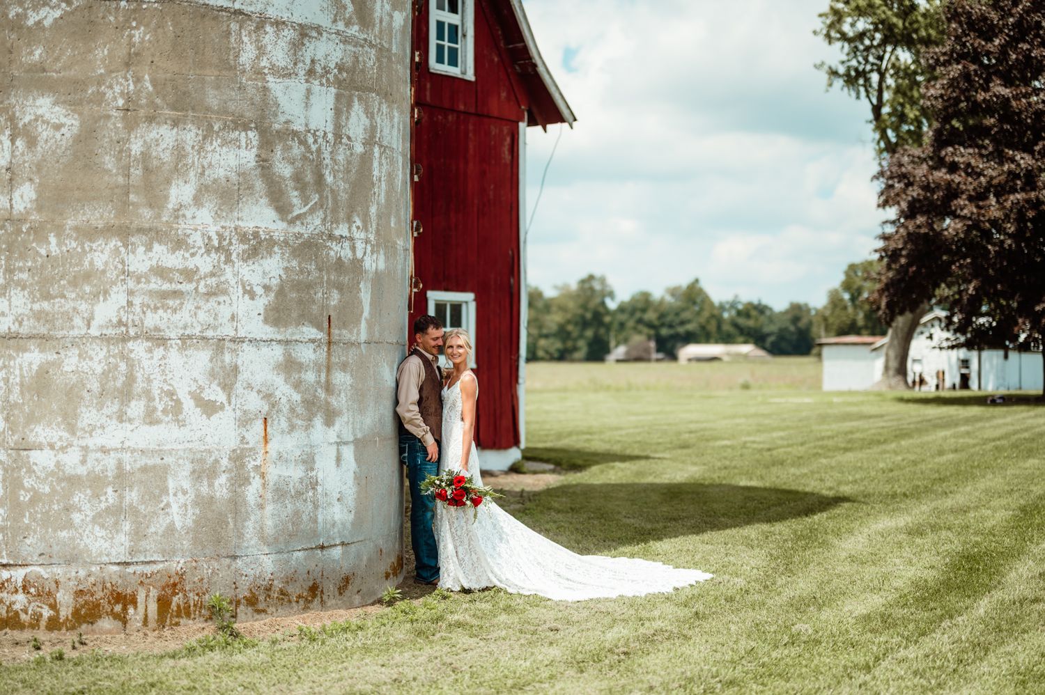 Bride and groom posing beside a rustic red barn on a lush green farm on their wedding day.