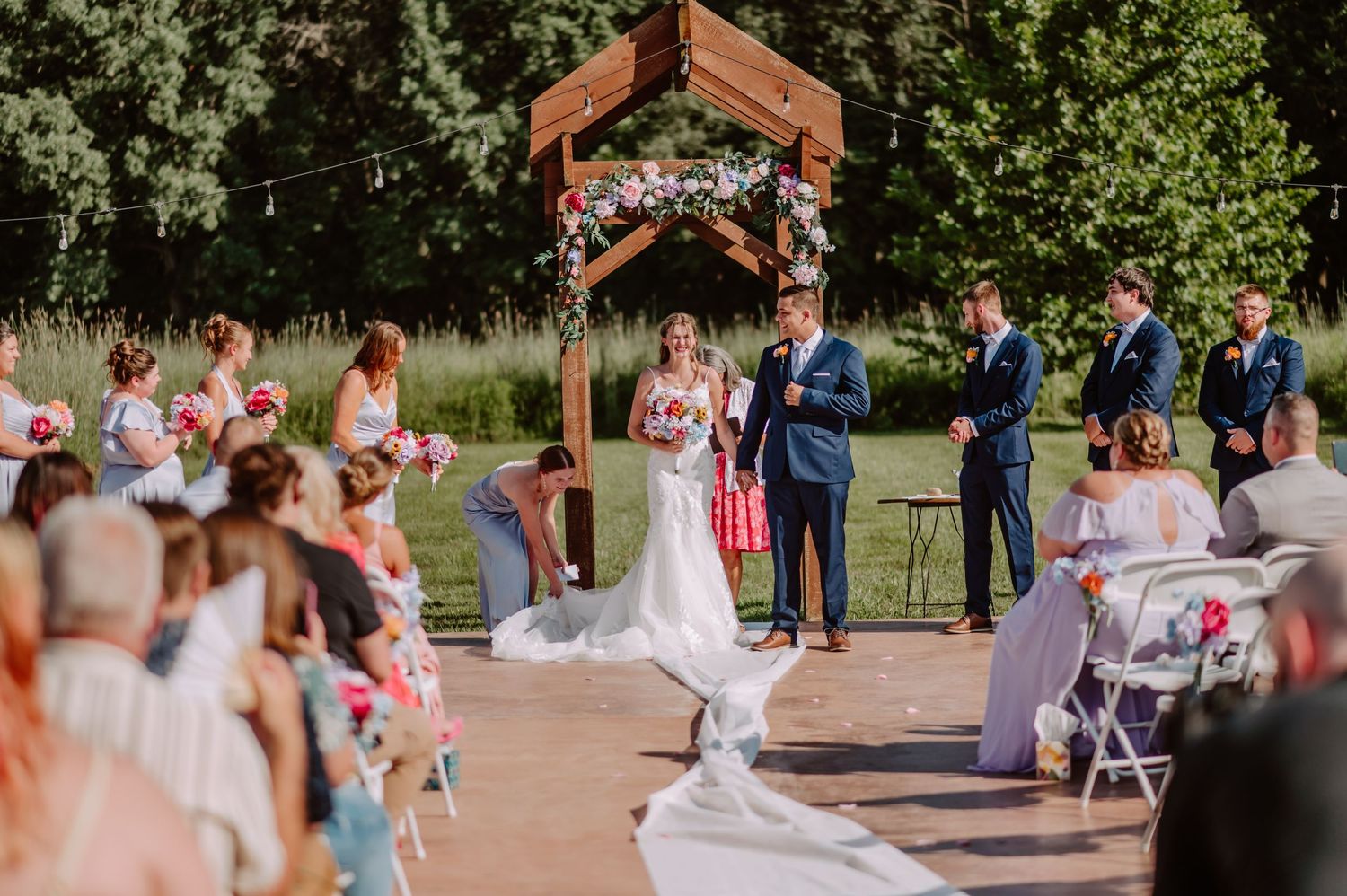 Bride walks down aisle toward groom at outdoor wedding ceremony under rustic wooden floral arch.