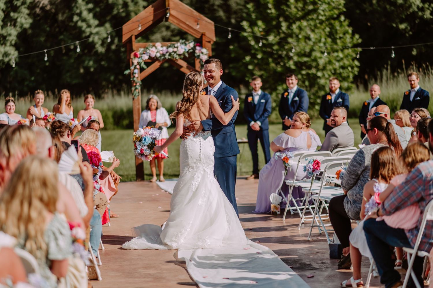 Newlyweds embrace and dance at outdoor wedding ceremony under string lights and floral arch.