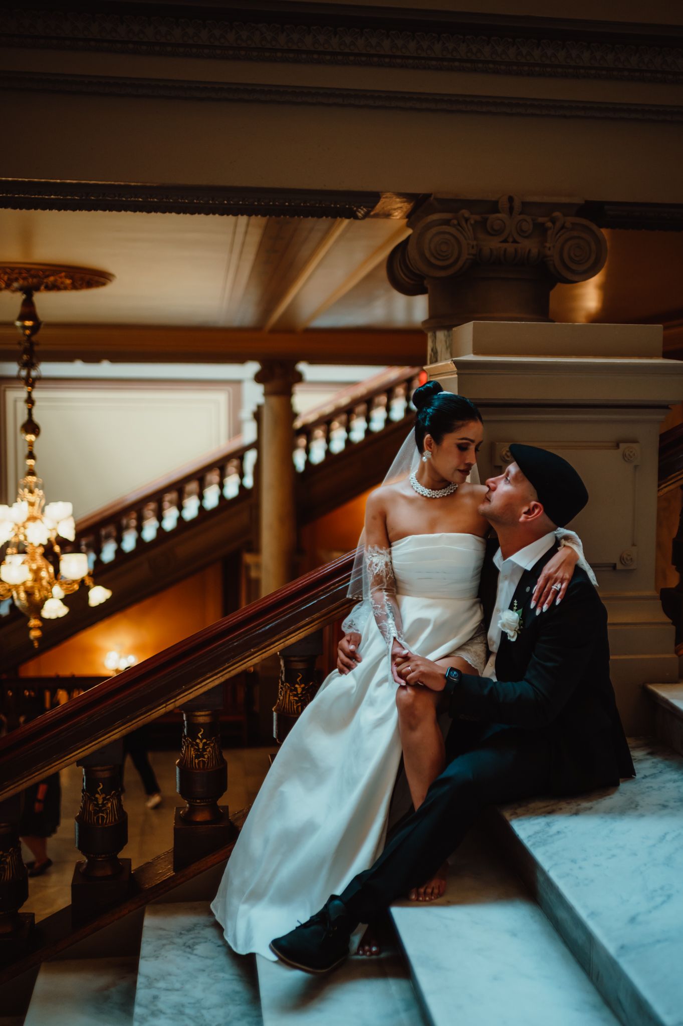 Bride and groom sit intimately on marble stairs in an elegant venue with chandeliers and ornate railings.