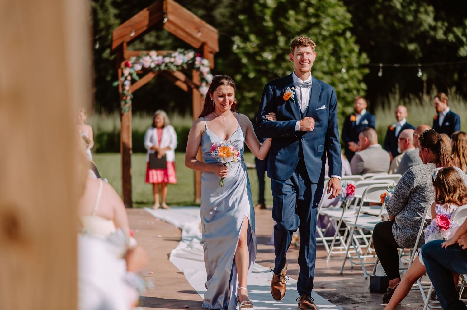 Bridesmaid in blue dress walks down outdoor wedding aisle with groomsman in navy suit past seated guests.
