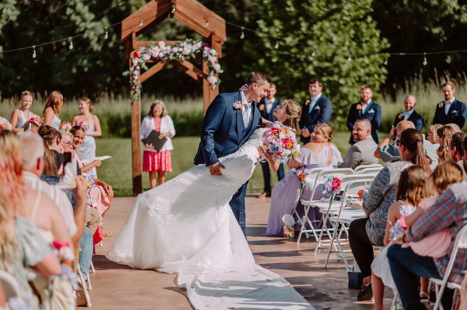 Groom dips bride for romantic kiss during outdoor wedding recessional with floral arch backdrop.