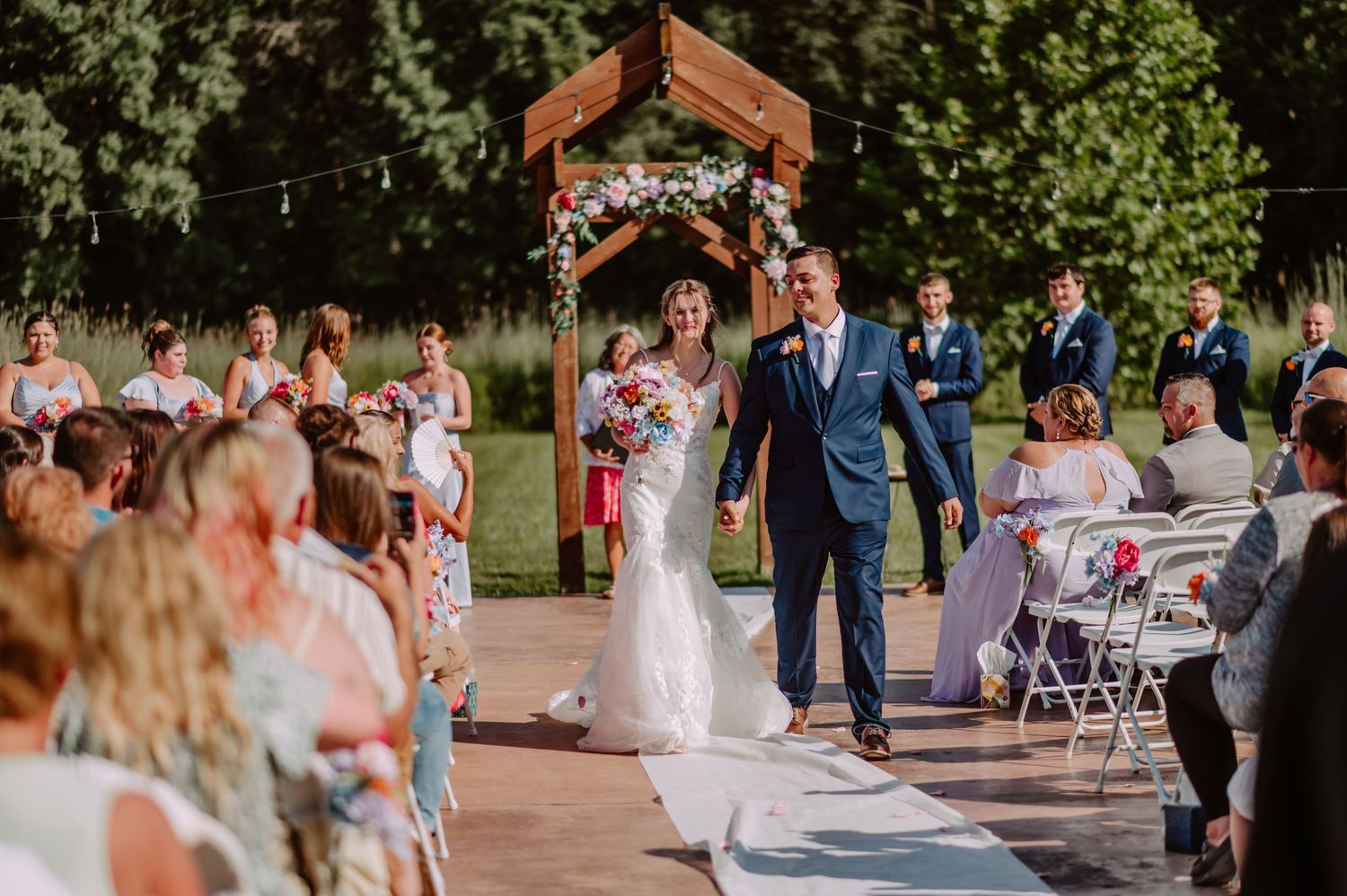 Bride and groom walk joyfully down the aisle after outdoor wedding ceremony under floral arch.