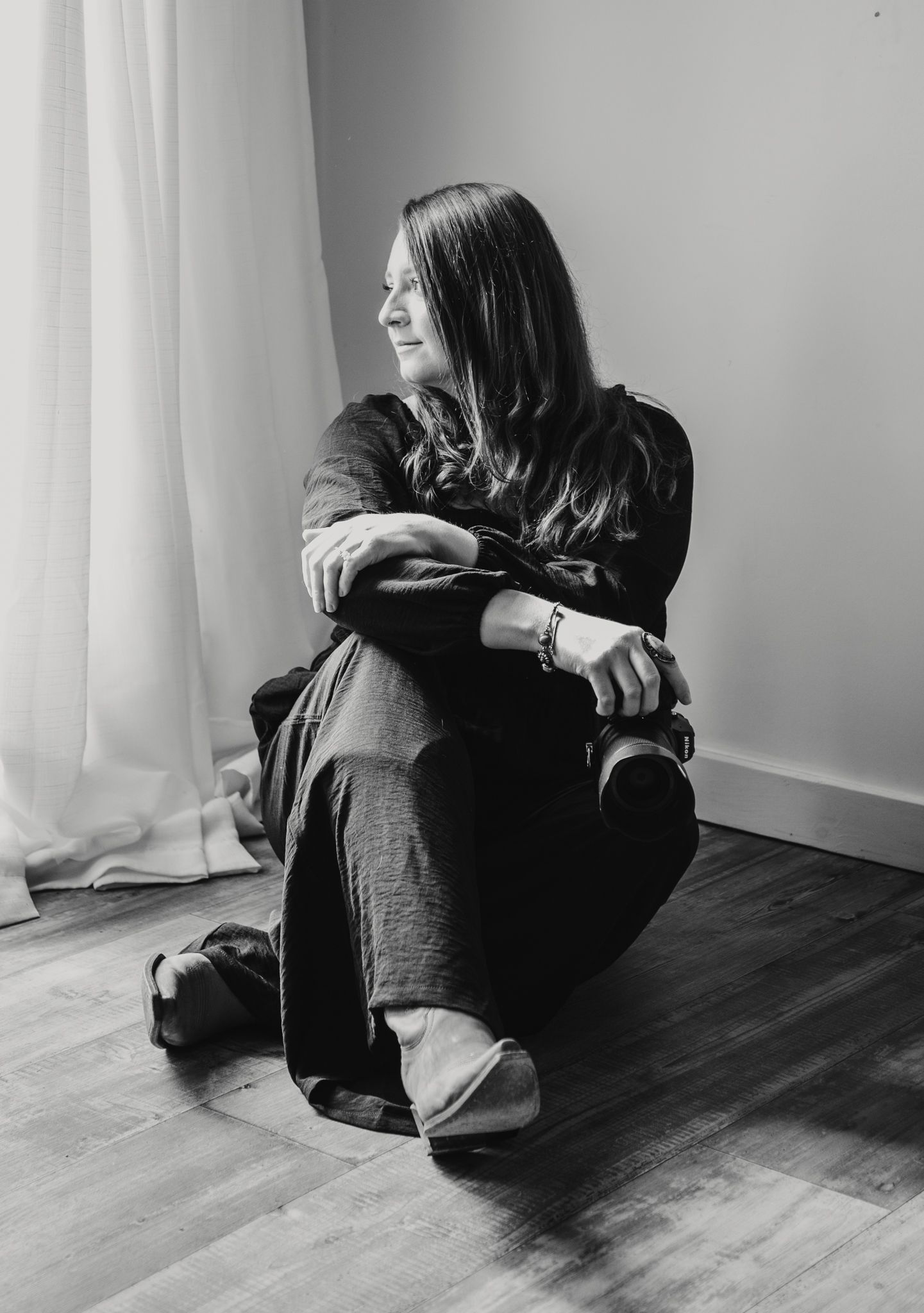 Black and white portrait of a female photographer sitting on a hardwood floor smiling near a curtained window.