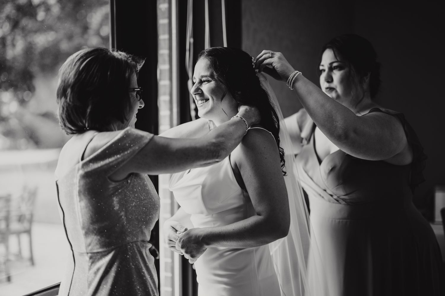 Black and white photo of a bride smiling as two women help her with her wedding dress and jewelry.