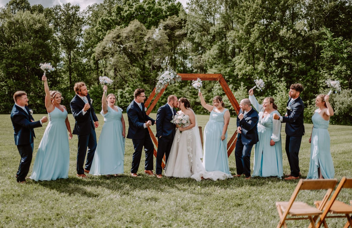Wedding party in blue dresses and navy suits celebrating outdoors with a wooden arch backdrop.