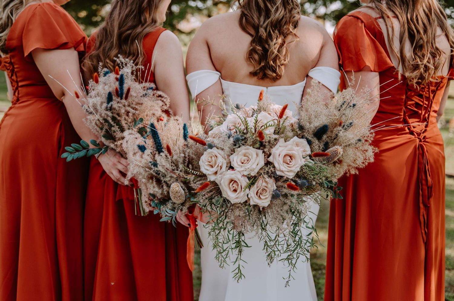 Bride in white gown with bridesmaids in rust red dresses holding boho floral bouquets with dried pampas grass.