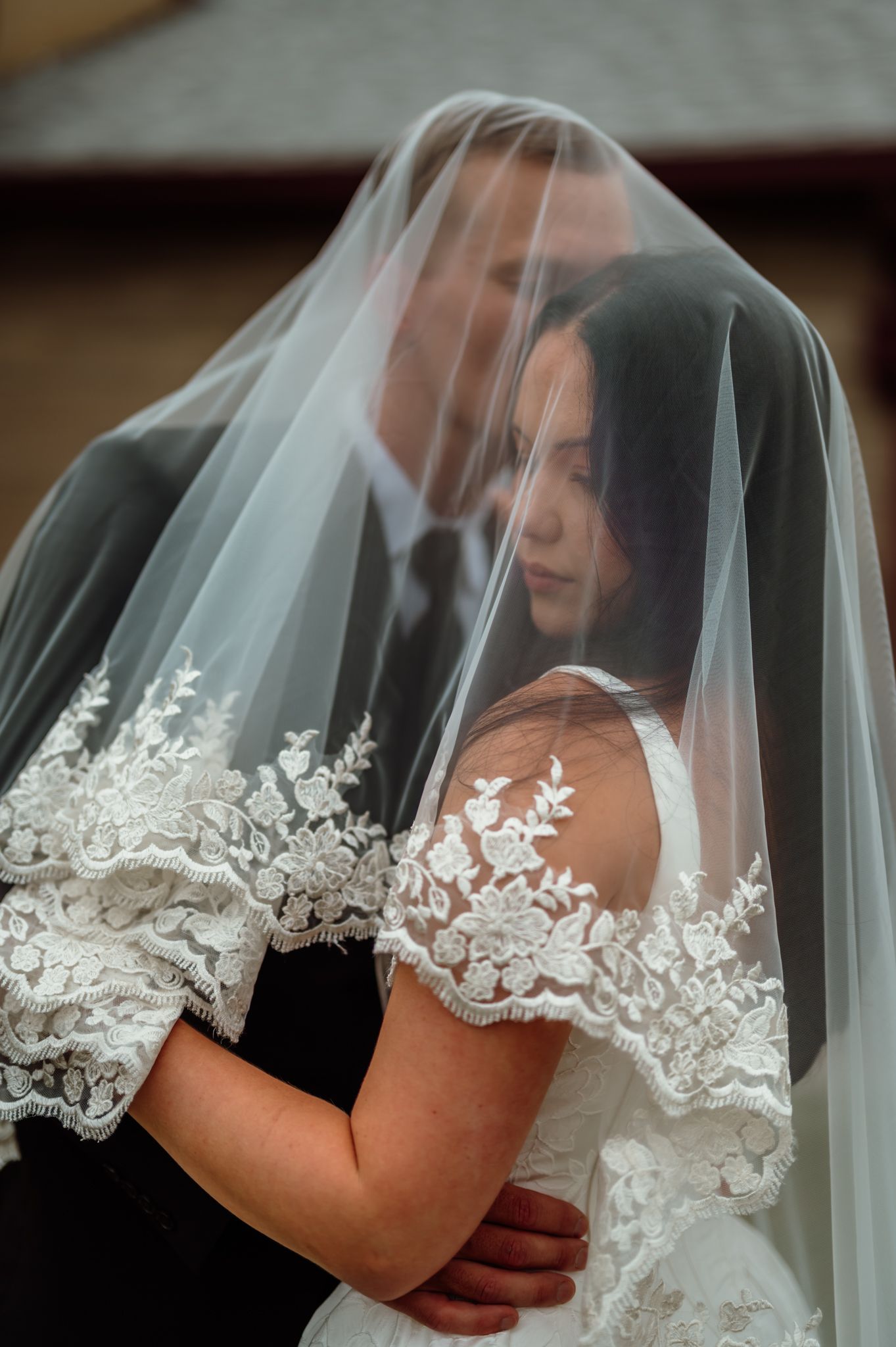 Bride and groom share intimate moment under lace-trimmed wedding veil, bride in elegant white gown embracing groom in black suit.