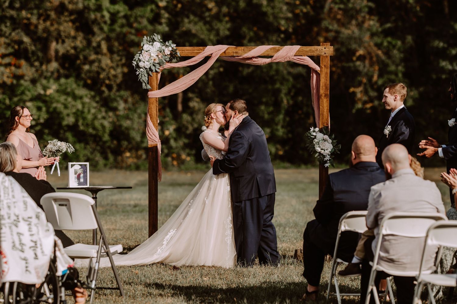 Bride and groom sharing first kiss under wooden arch at outdoor wedding ceremony with seated guests.
