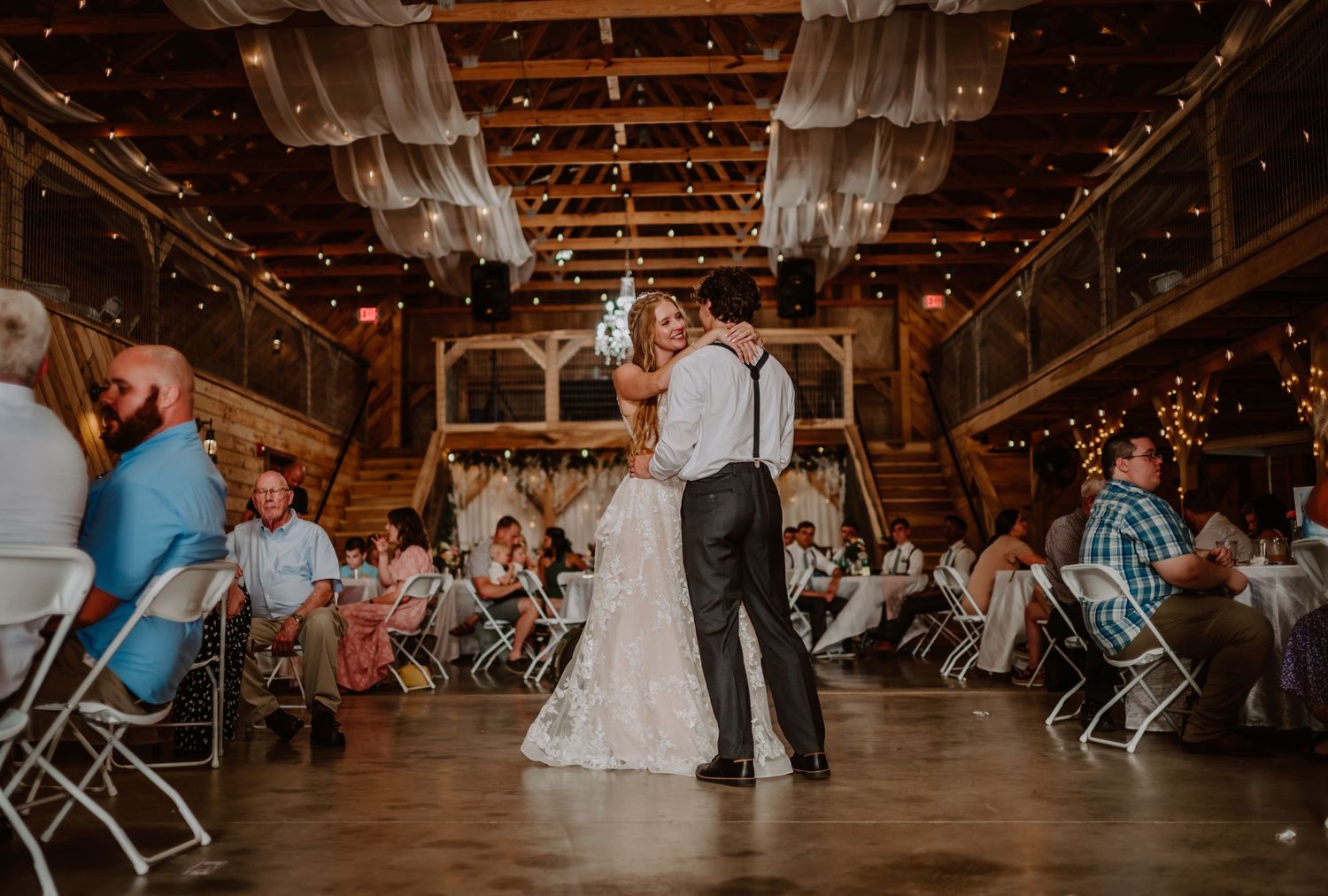 Newlyweds share romantic first dance kiss in elegant barn reception venue with guests seated.