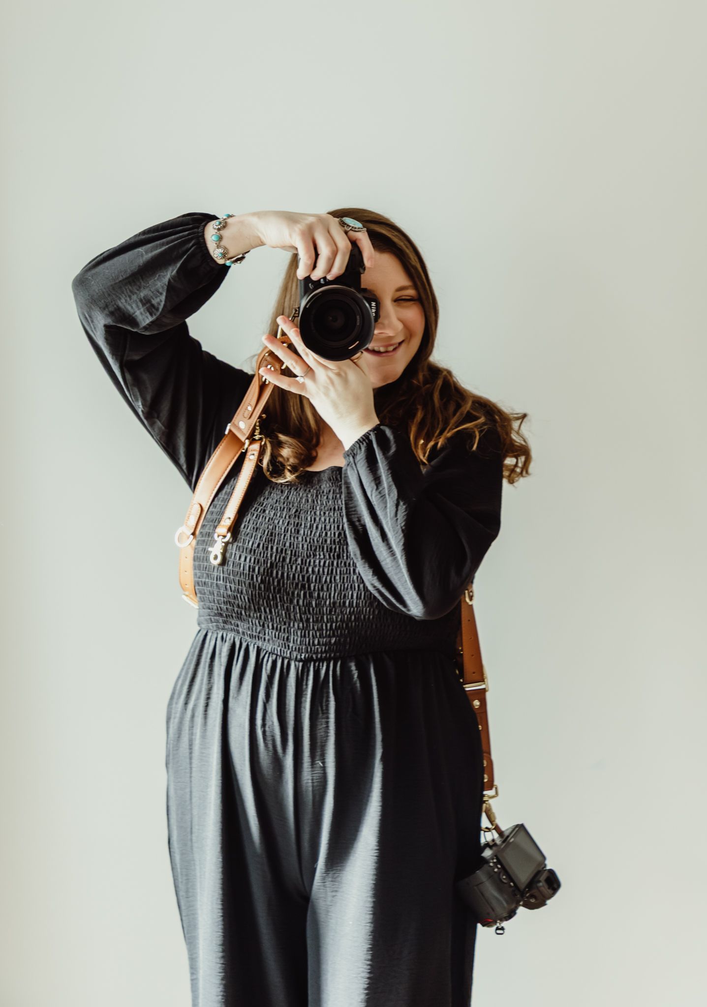 Smiling female photographer in black dress shooting with DSLR camera and leather camera strap against white background.