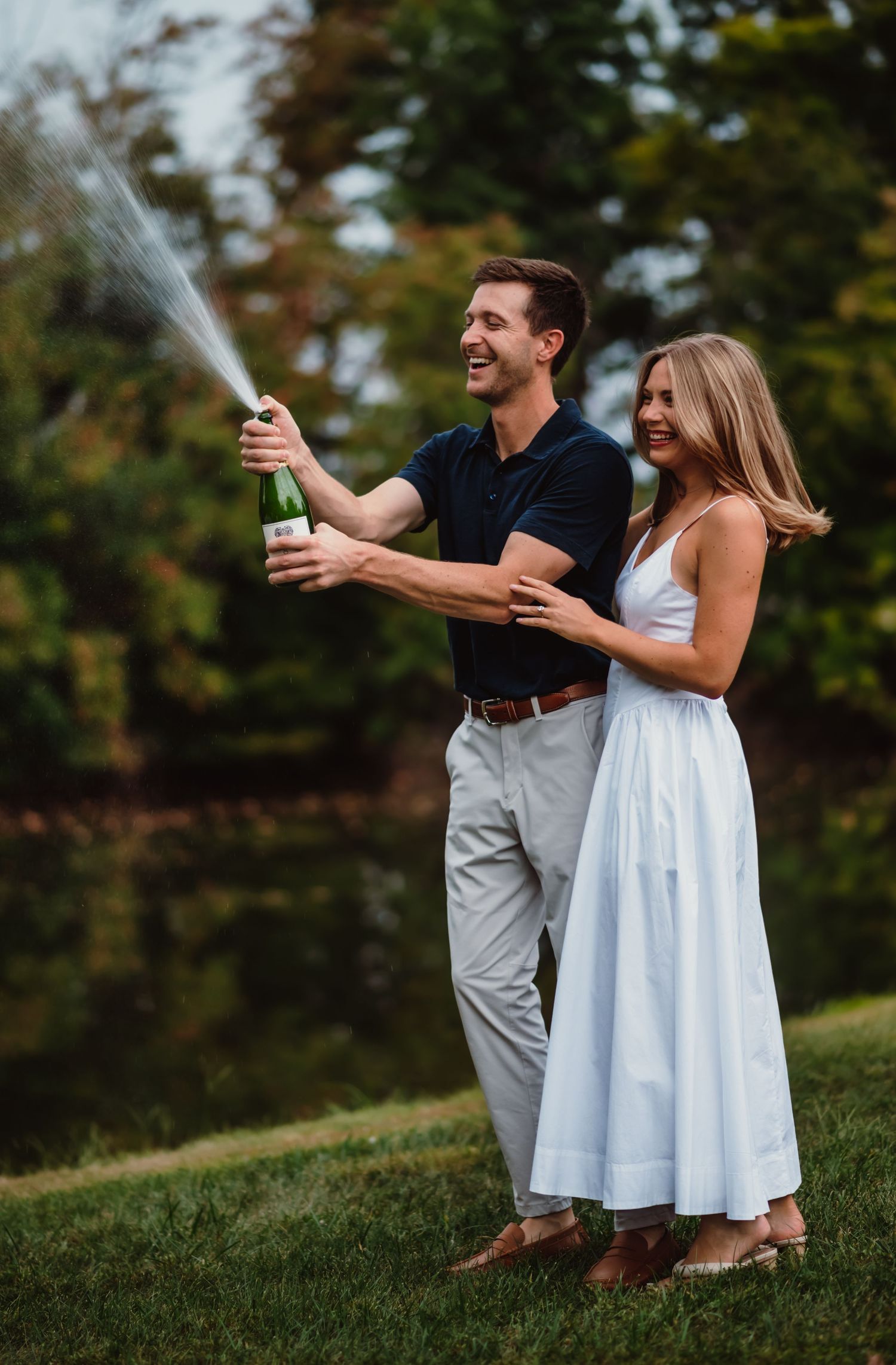 Happy couple celebrating outdoors, popping champagne bottle, woman in white dress, man in navy polo, lush green park background.
