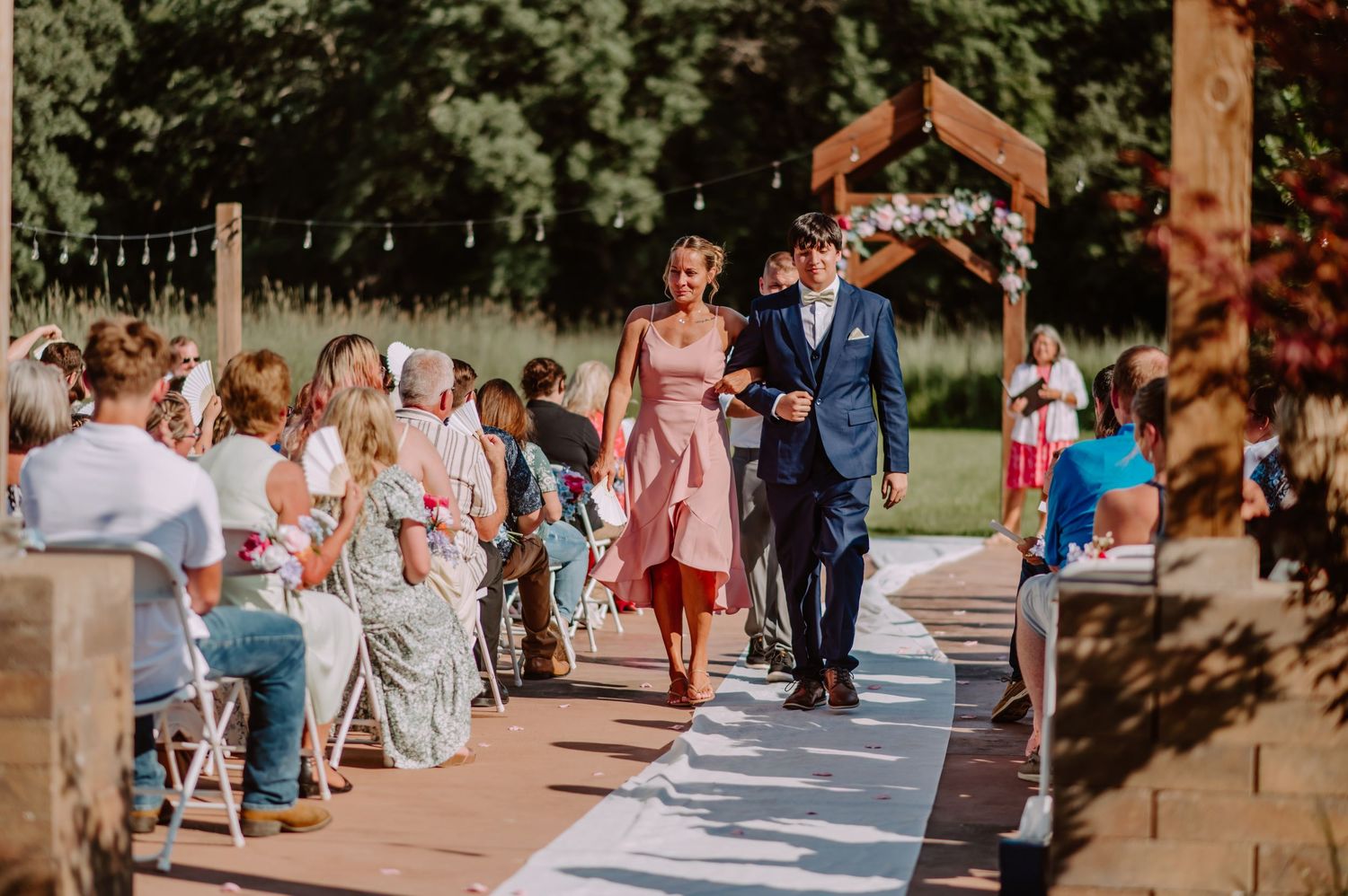 Bridesmaid in pink dress walks down outdoor wedding aisle escorted by groomsman in navy suit.