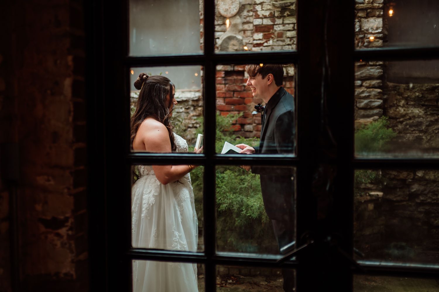Bride and groom exchange vows through a rustic window at a charming stone venue wedding ceremony.