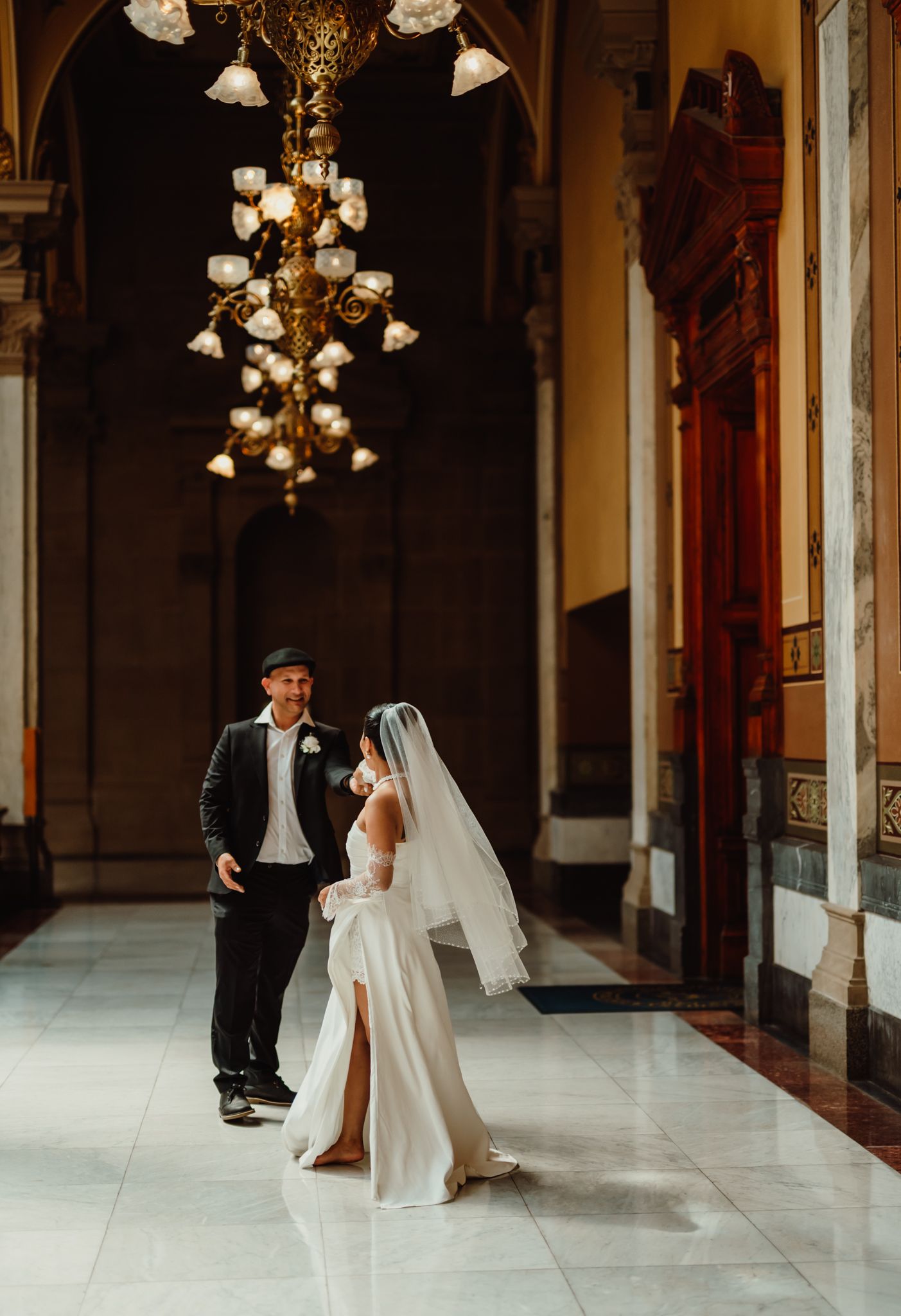Bride in white gown with veil and groom in black suit share a moment in an elegant chandelier-lit marble hall.