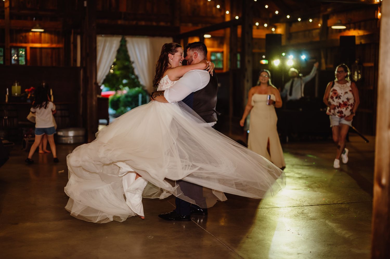 Groom lifting bride in elegant white gown during first dance in rustic barn wedding venue with string lights.