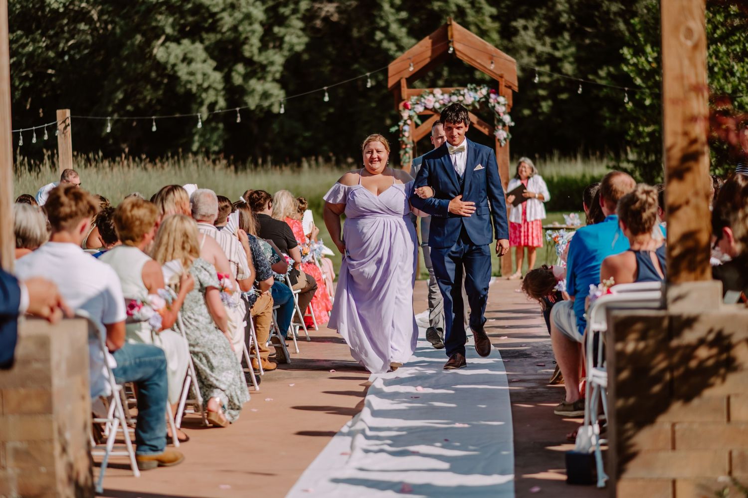 Groomsman in navy suit escorts woman in lavender dress down outdoor wedding aisle past seated guests.