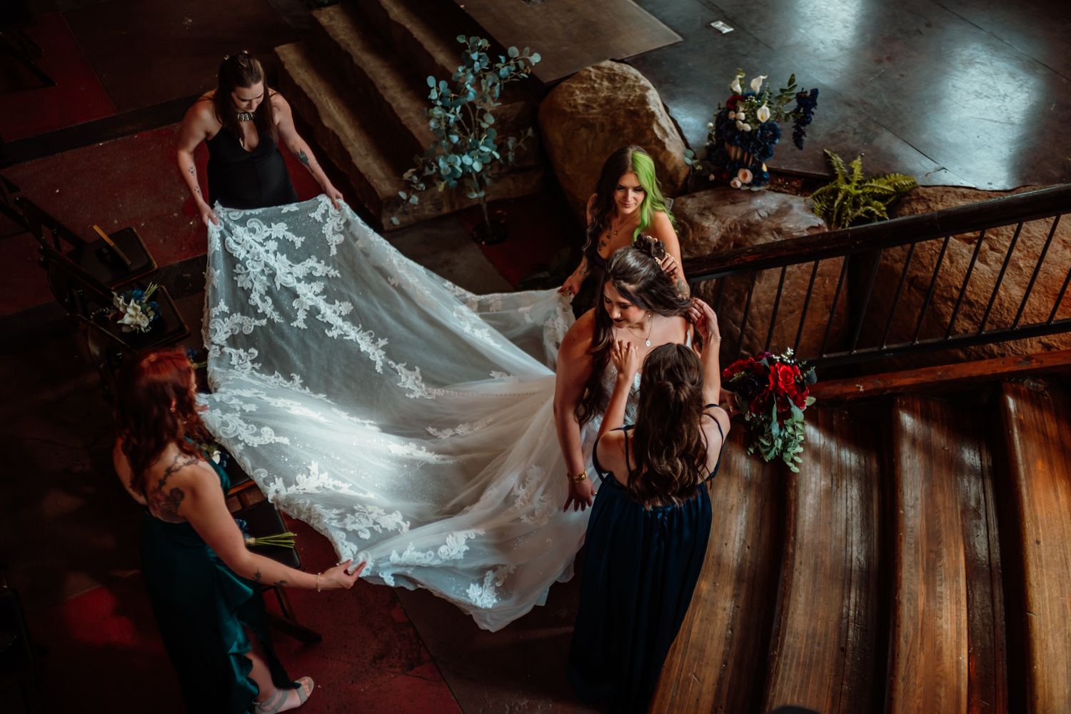 Aerial view of bride in white lace gown with bridesmaids adjusting her train on ornate wooden staircase.