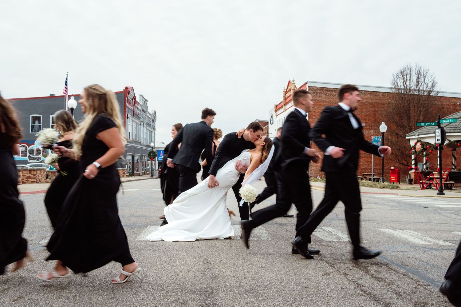Bride and groom share a romantic dip kiss while wedding party walks across a downtown street in formal attire.