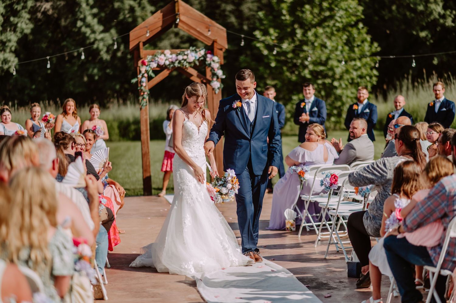 Happy newlyweds walk down aisle at outdoor wedding ceremony surrounded by cheering guests.