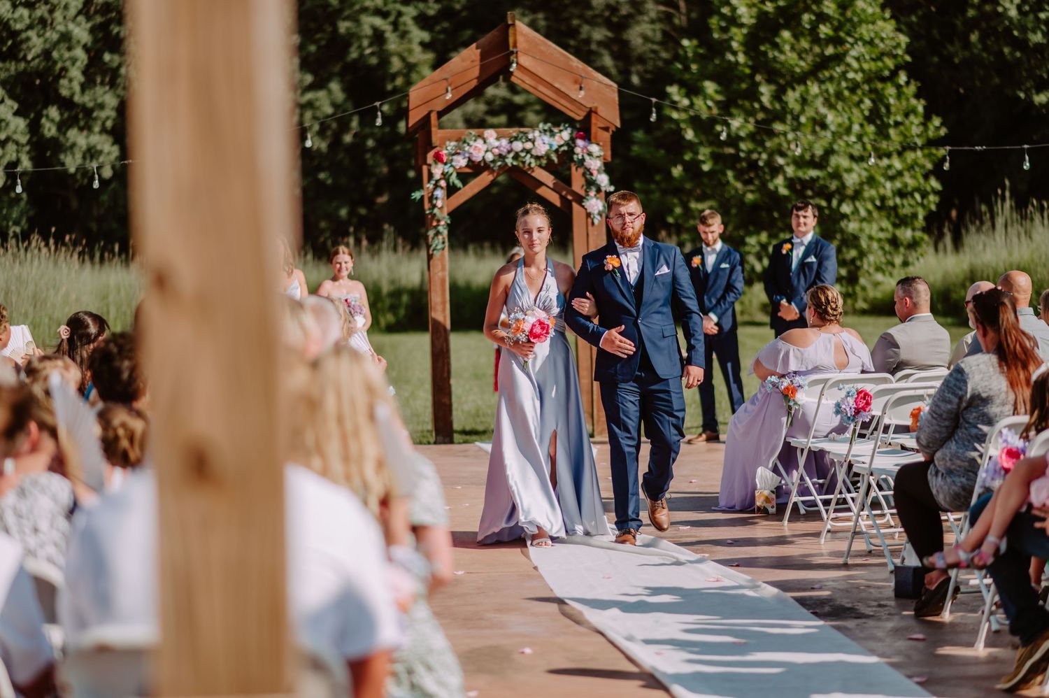 Wedding party processional with bridesmaid and groomsman walking down aisle toward wooden floral arch.