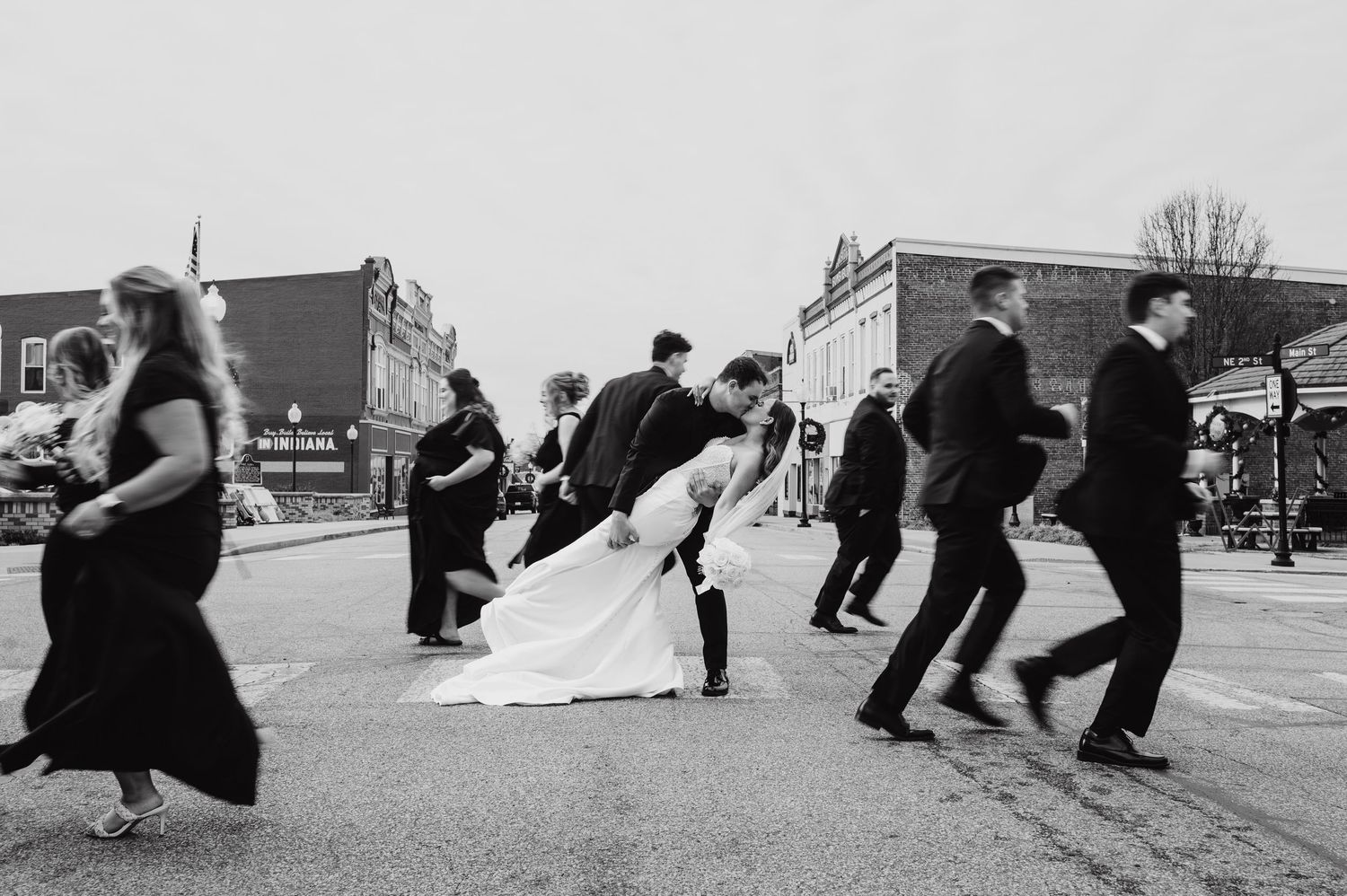 Black and white photo of bride and groom sharing a dip kiss while wedding party crosses a small town street.