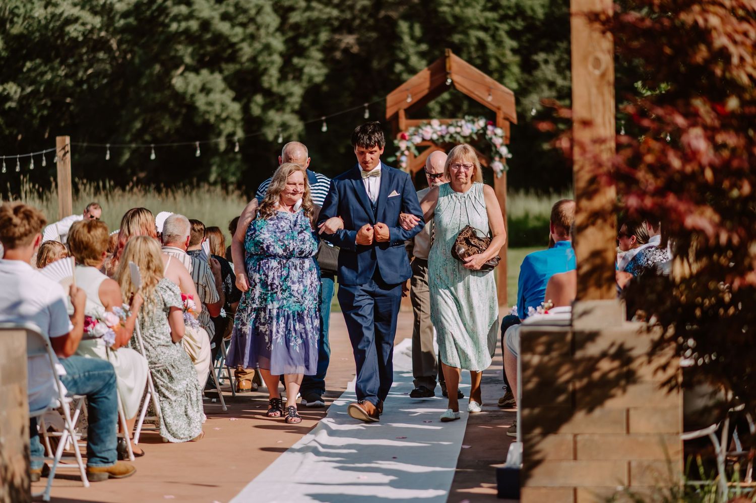 Groom in navy suit is escorted down the aisle by two women at an outdoor wedding ceremony.