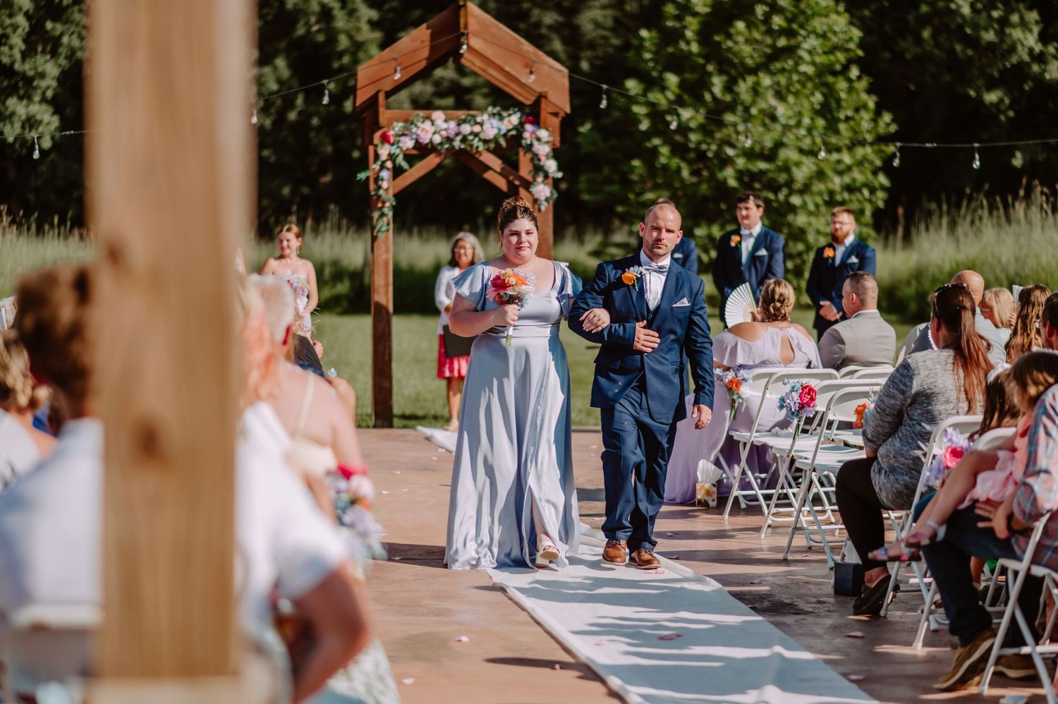 Bridesmaid in periwinkle dress and groomsman in navy suit walk down outdoor wedding ceremony aisle.
