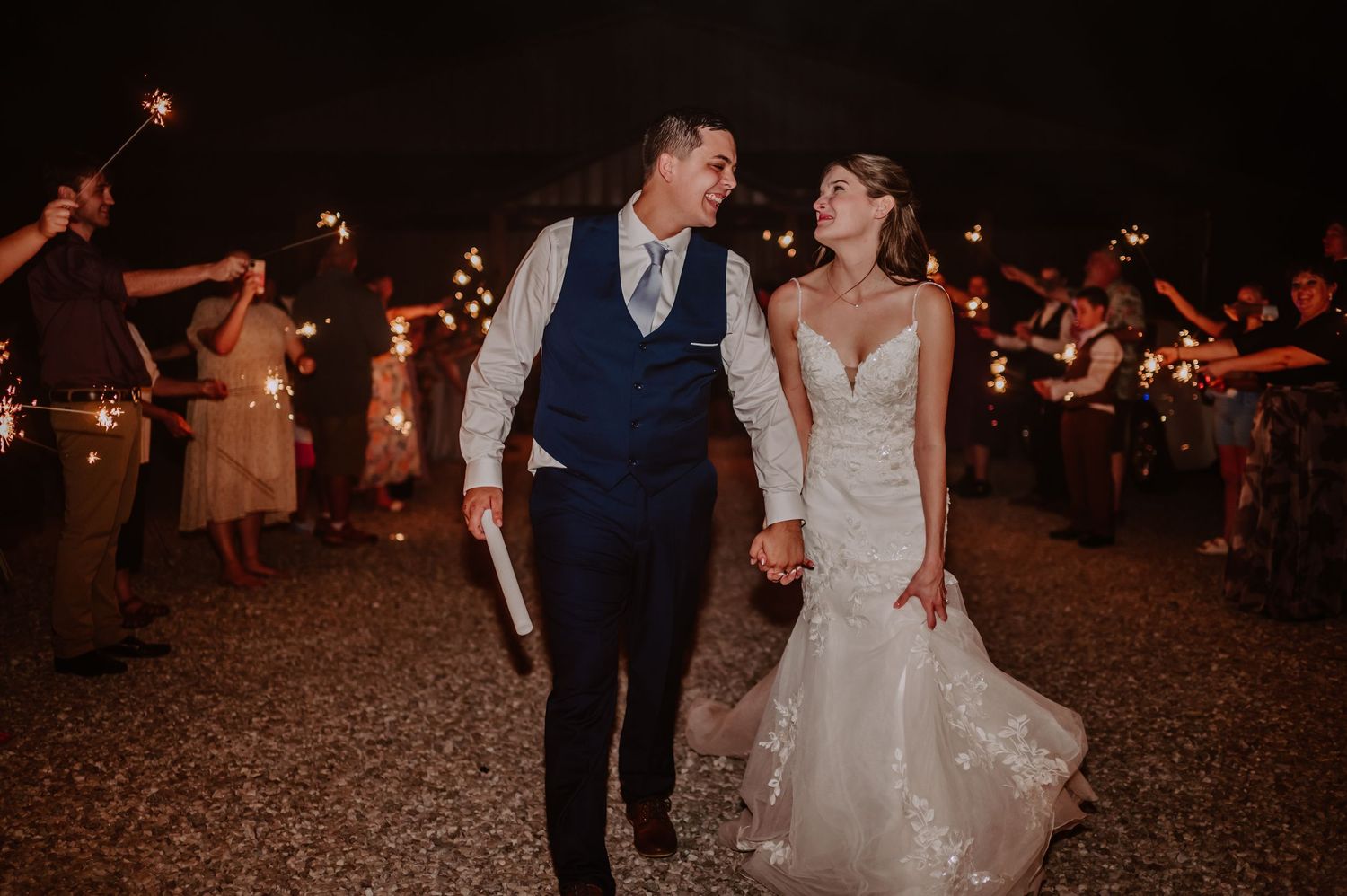 Bride and groom smile during sparkler exit at night surrounded by cheering wedding guests.
