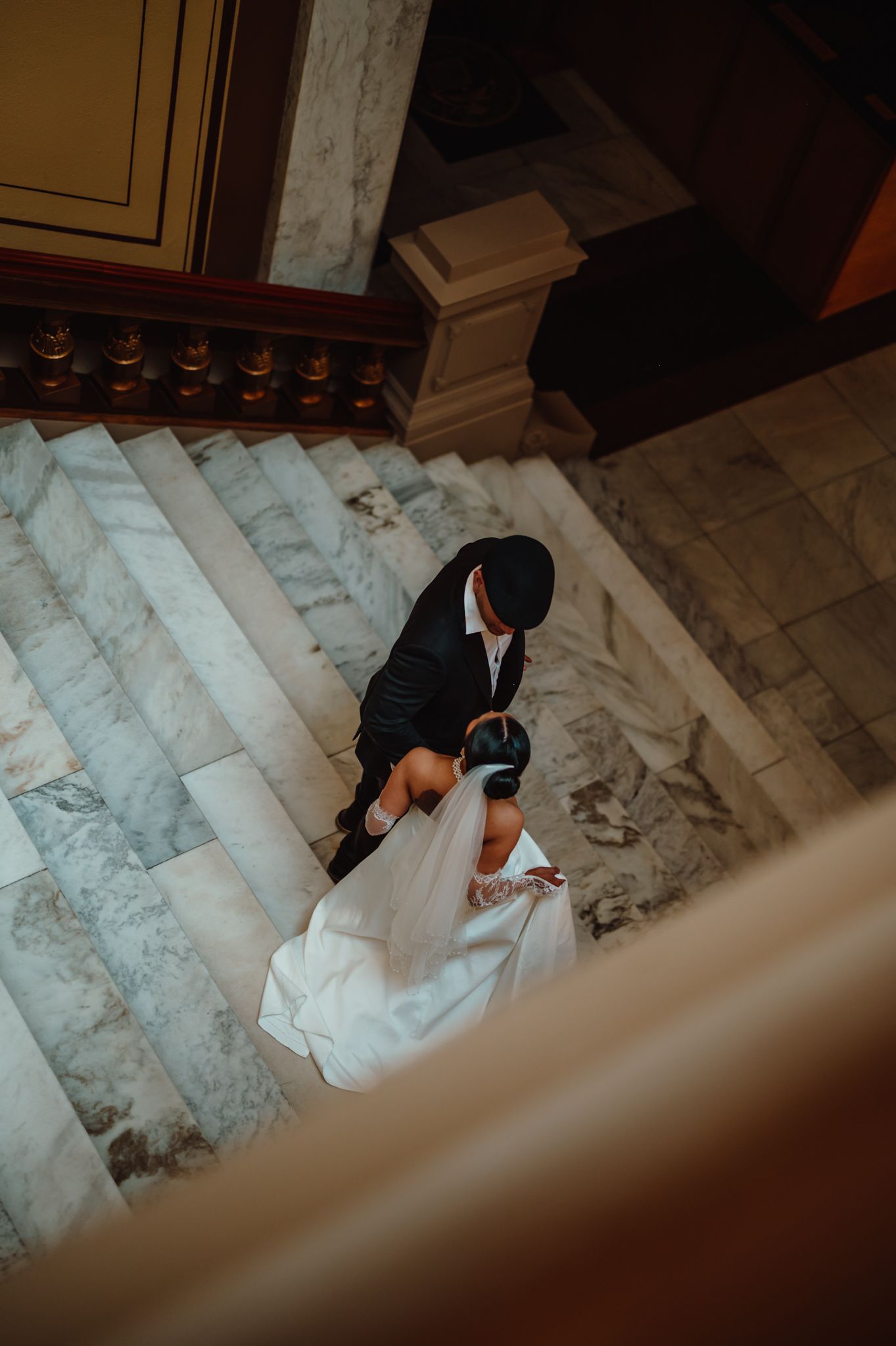 Aerial view of a bride and groom sharing a romantic moment on elegant marble stairs in a grand venue.