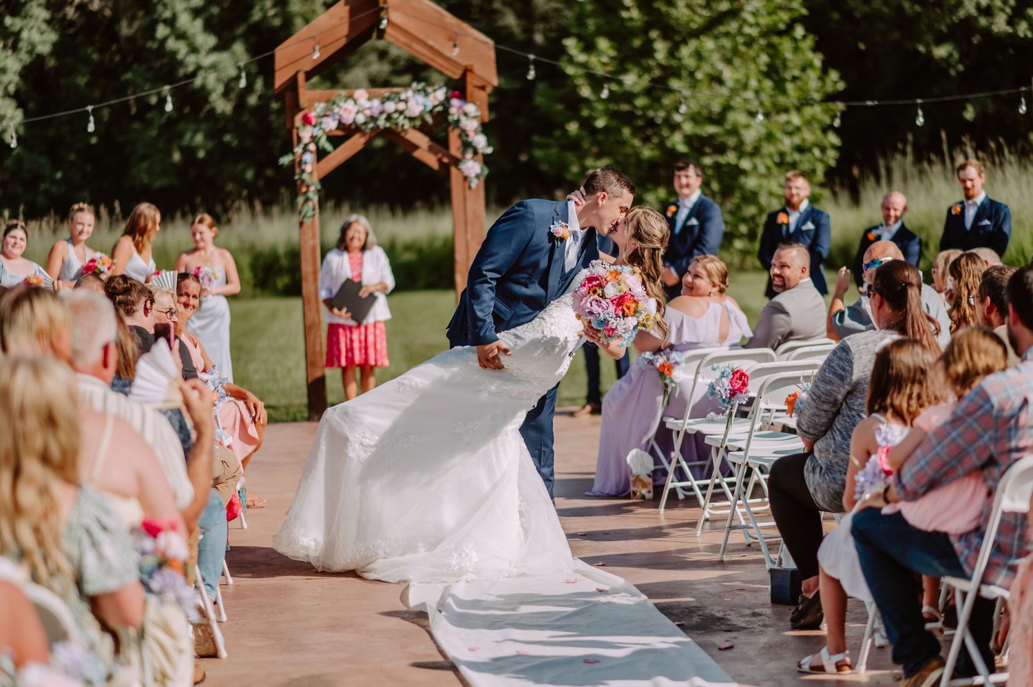 Groom dips bride for kiss during outdoor wedding recessional while guests cheer and celebrate.