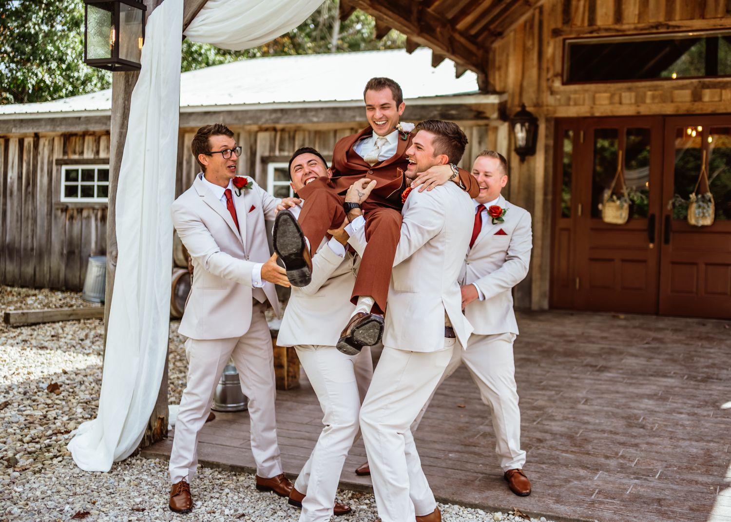 Groomsmen joyfully lift the groom at a rustic outdoor wedding venue The Barn at Timber Ridge. Indianapolis wedding photographer