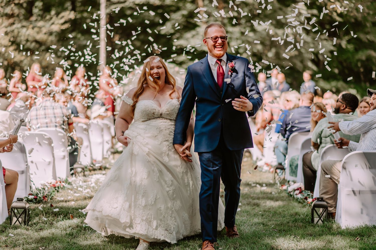 Bride and groom walk down outdoor aisle as guests toss white flower petals during sunny wedding ceremony. Indianapolis wedding photographer