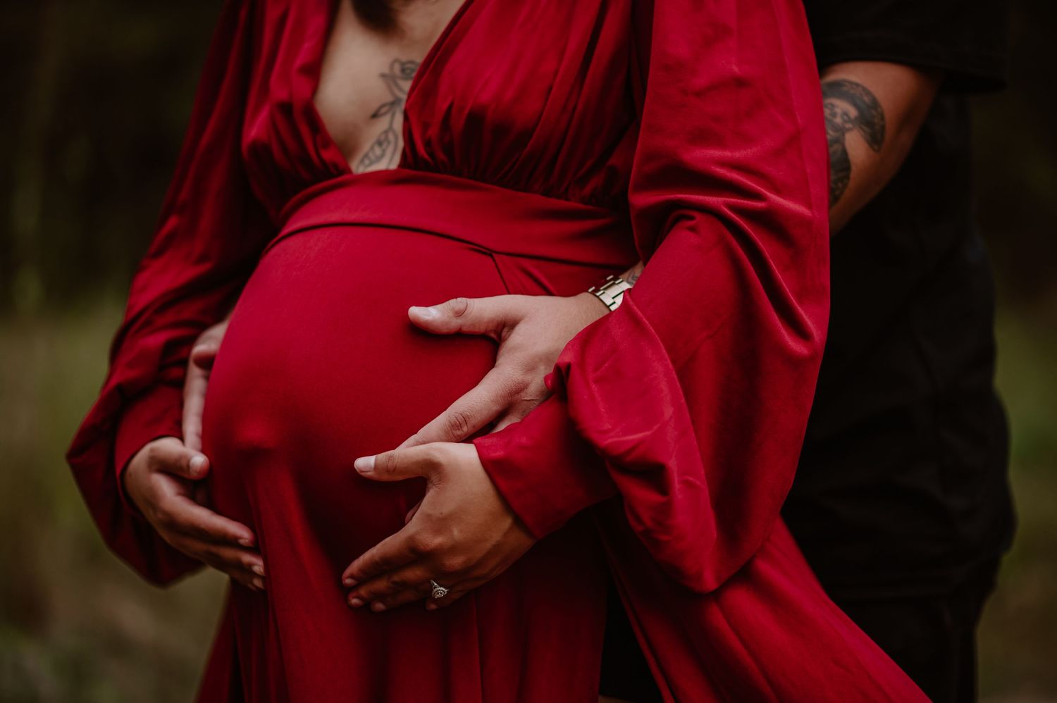 Pregnant woman in red dress cradling baby bump with partner's tattooed hand, maternity photo shoot outdoors.