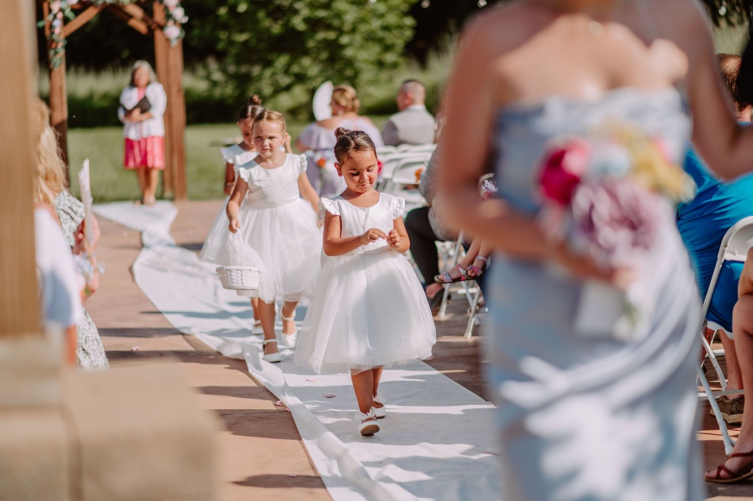 Two adorable flower girls in white dresses smile while walking down outdoor wedding ceremony aisle.