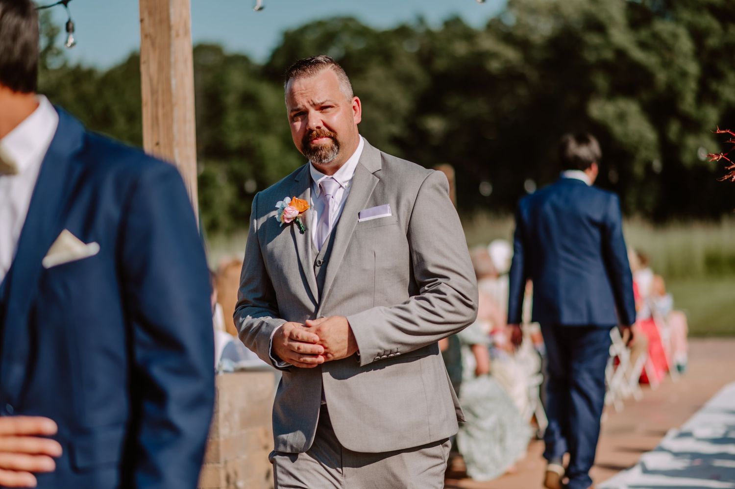 Groomsman in gray suit with boutonniere stands attentively at an outdoor wedding ceremony.