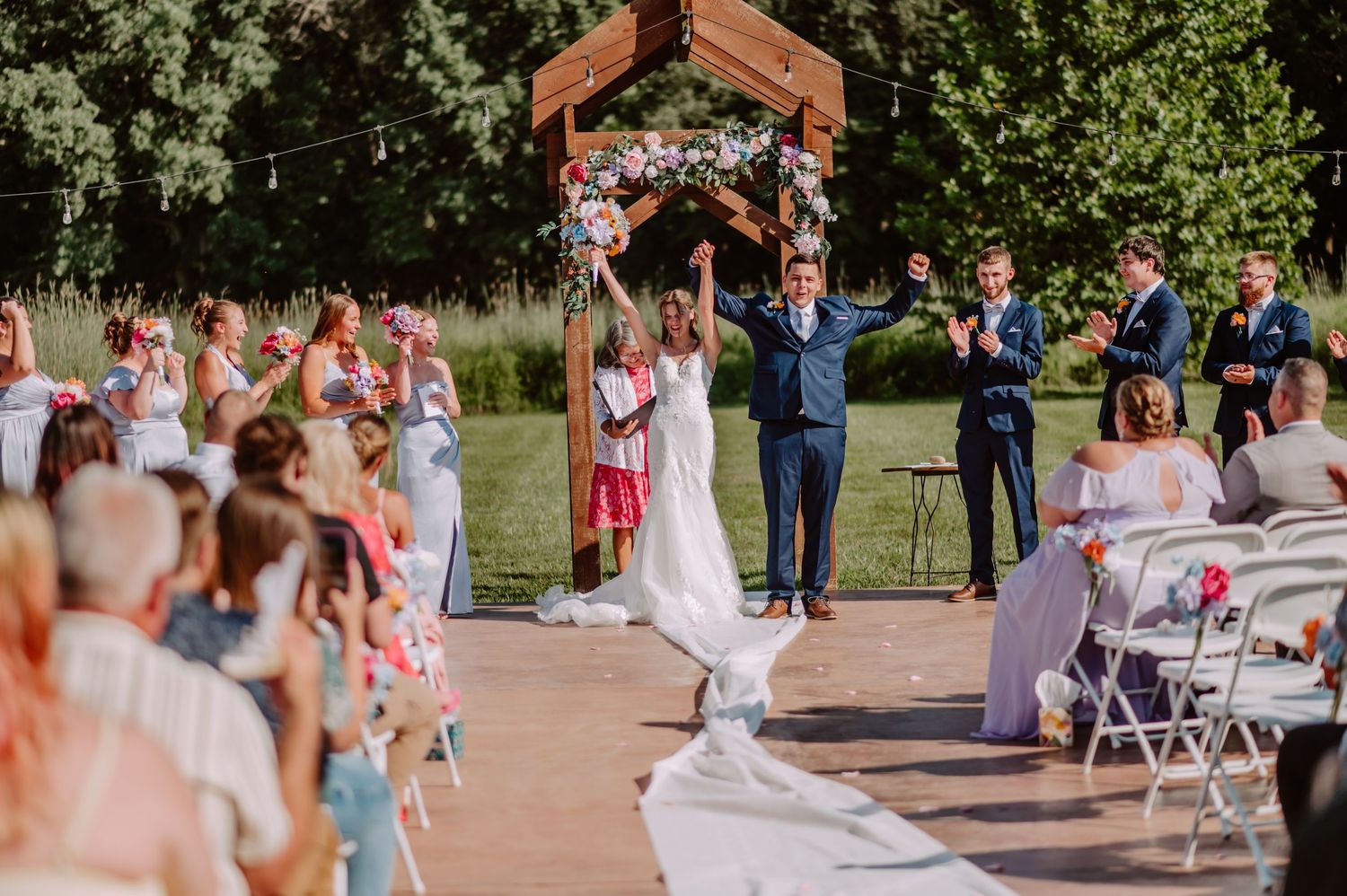 Newlyweds raise hands in celebration under wooden floral arch after outdoor wedding ceremony.