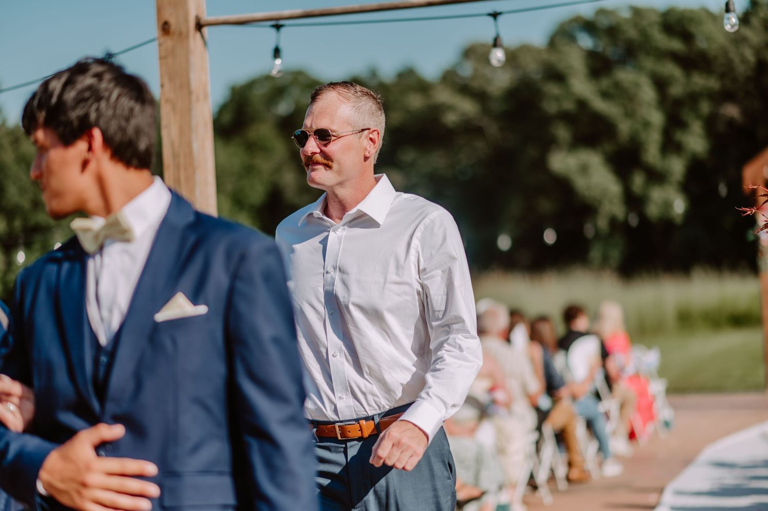 Man in white shirt and sunglasses smiling at an outdoor wedding ceremony.