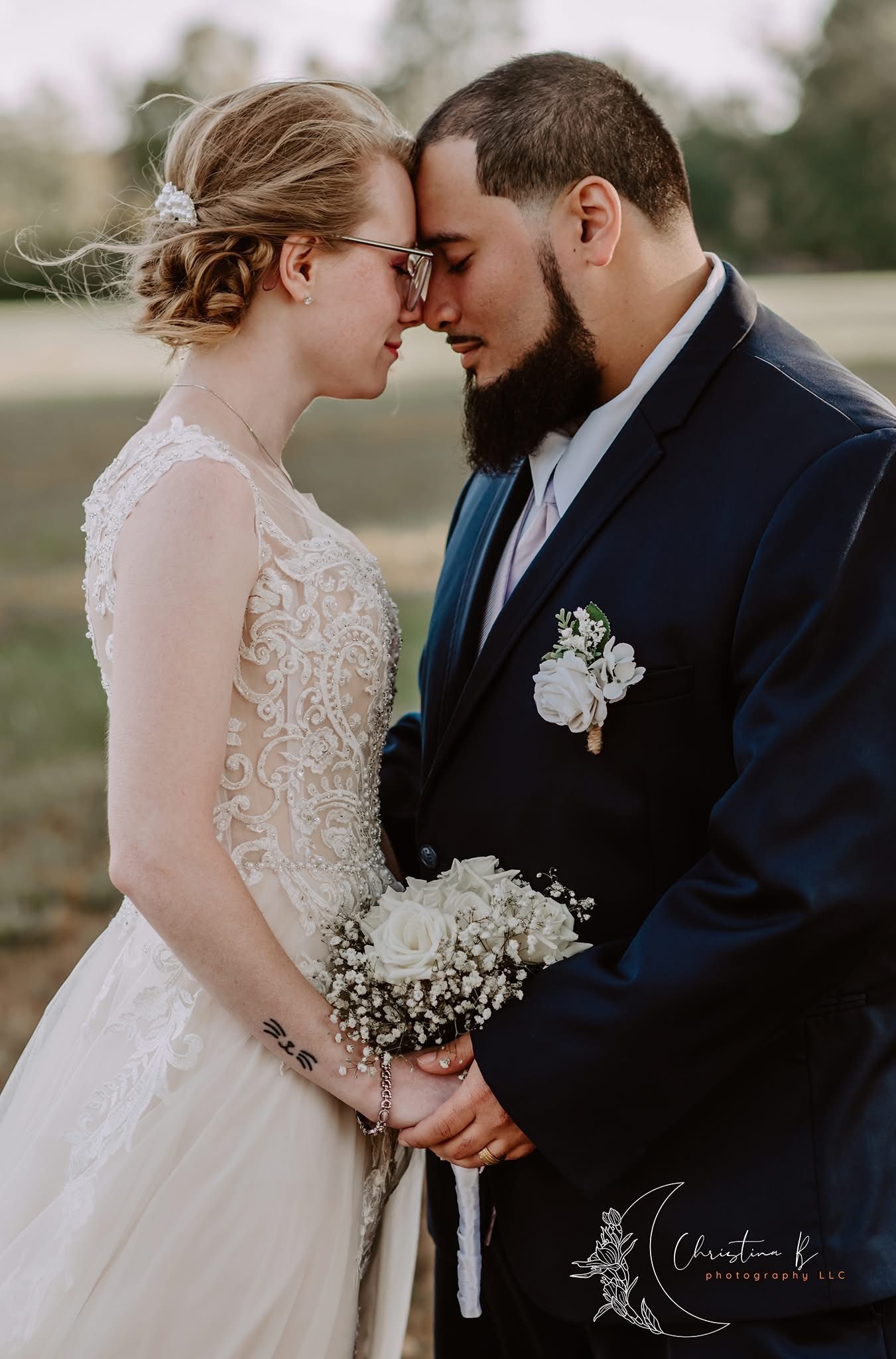 Bride and groom share an intimate forehead touch moment, bride holding white bouquet in lace gown, groom in navy suit.
