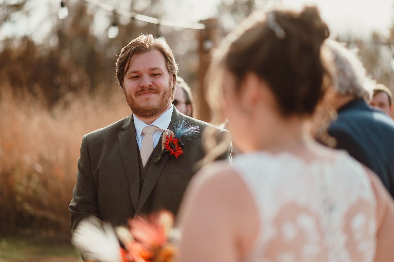 Groom smiling warmly at bride during outdoor wedding ceremony with golden hour lighting and red boutonniere.