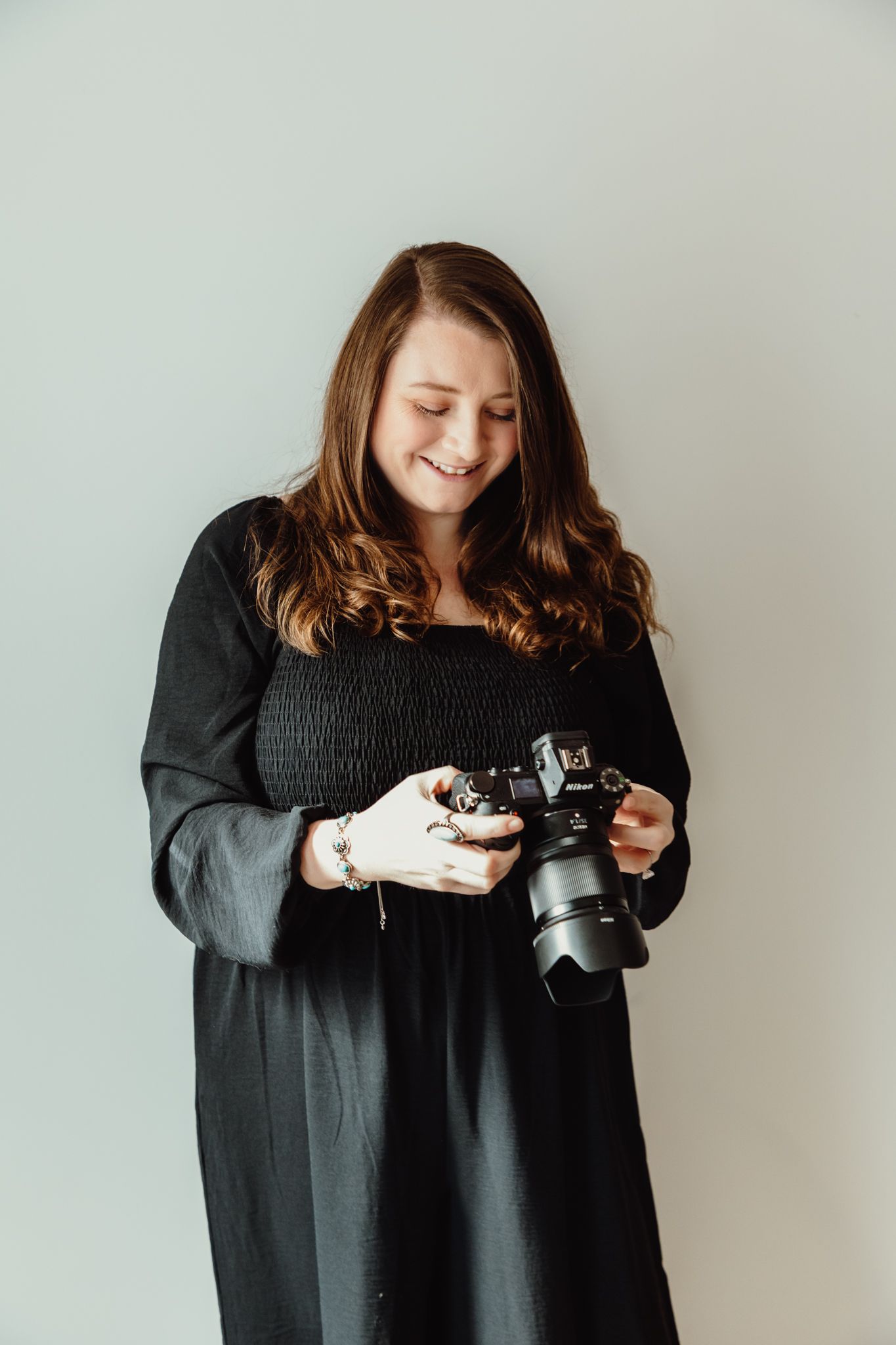 Happy female photographer in black dress looking down at DSLR camera screen against a light gray background.