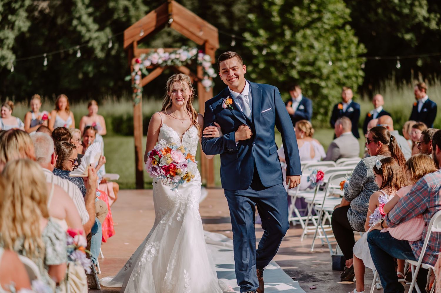 Excited bride holding colorful bouquet and groom in navy suit celebrate recessional at outdoor wedding.