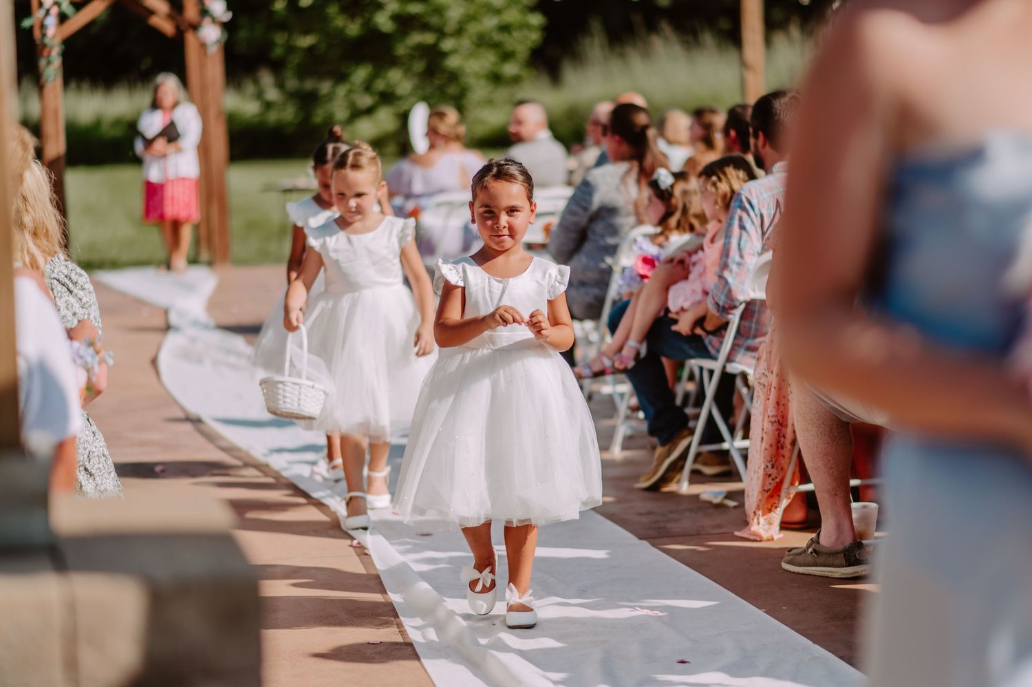 Adorable flower girls in white tulle dresses scatter petals along the aisle at an outdoor wedding ceremony.
